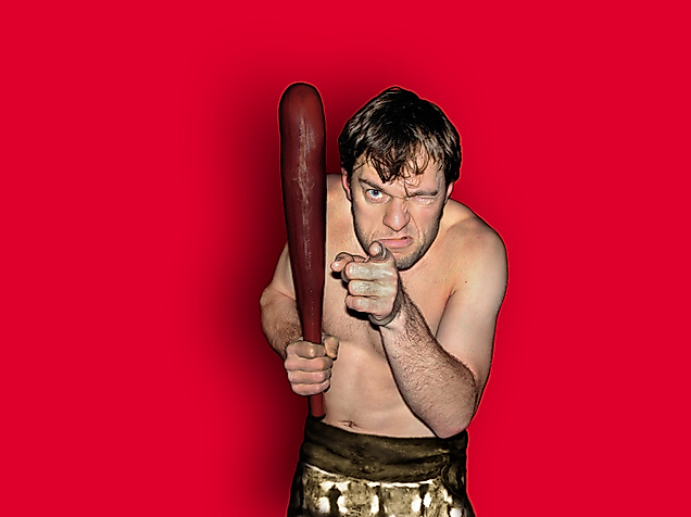 Szenenbild Cavemen: Ein Mann mit Beil, oberkörperfrei zeigt mit einem Finger in die Kamera und schaut direkt rein