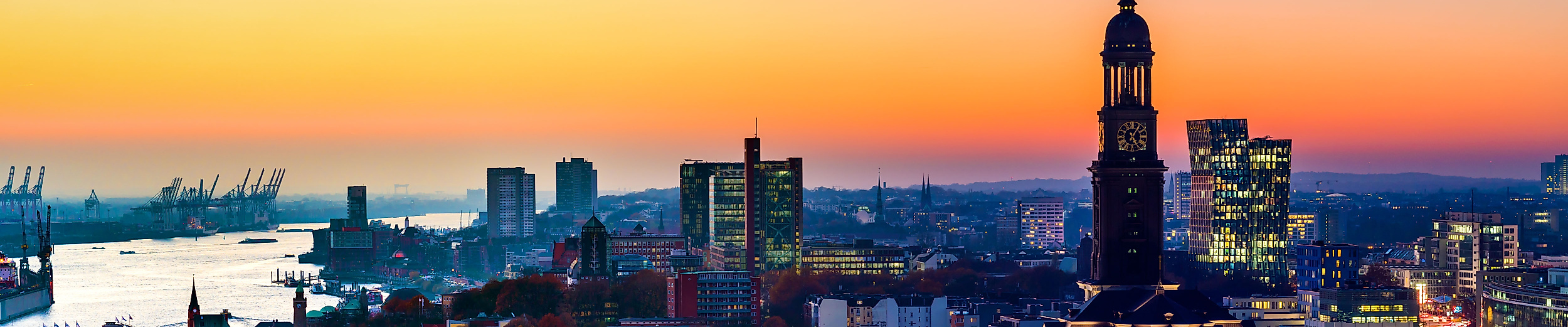 Panorama von Hamburg bei Sonnenuntergang mit beleuchtetem Michel-Turm und Hafen im Hintergrund.