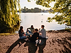 Gruppe beim Picknick am Alsterufer im Sommer mit Blick auf Boote und Sonne auf dem Wasser