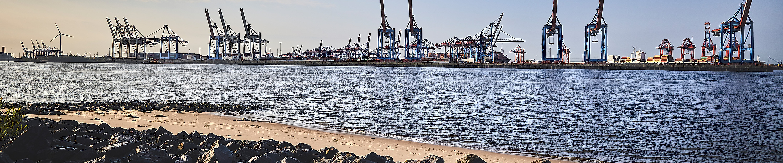 Blick vom Elbstrand auf Containerkräne im Hamburger Hafen bei Abendlicht, mit Steinen im Vordergrund