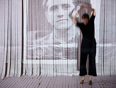 Die performende Person bewegt sich mit erhobenen Armen im Raum. Dahinter ist ein Vorhang aus weißen Streifen, auf den das Führerscheinbild eines Mannes zu sehen ist.