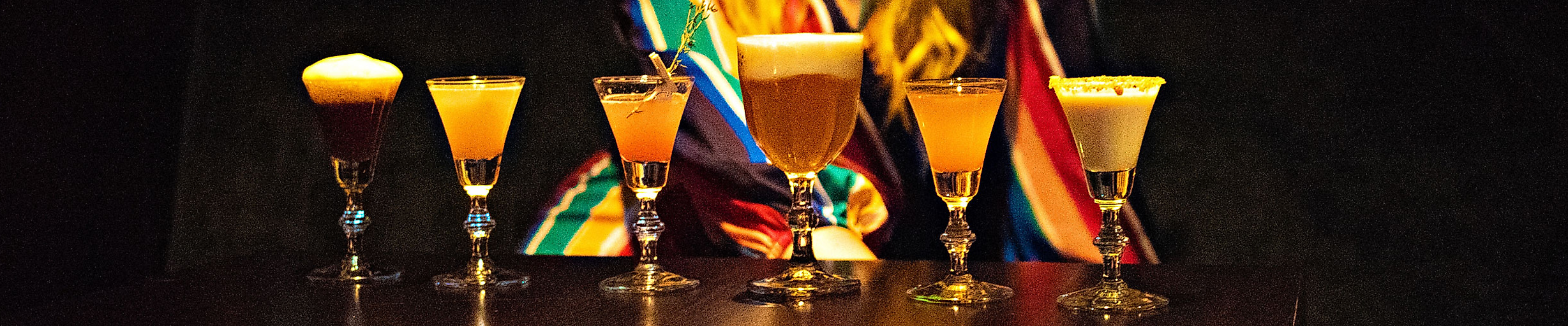 Sechs bunte Cocktails auf dunkler Theke, dahinter Person in bunter Kleidung vor Clubkulisse