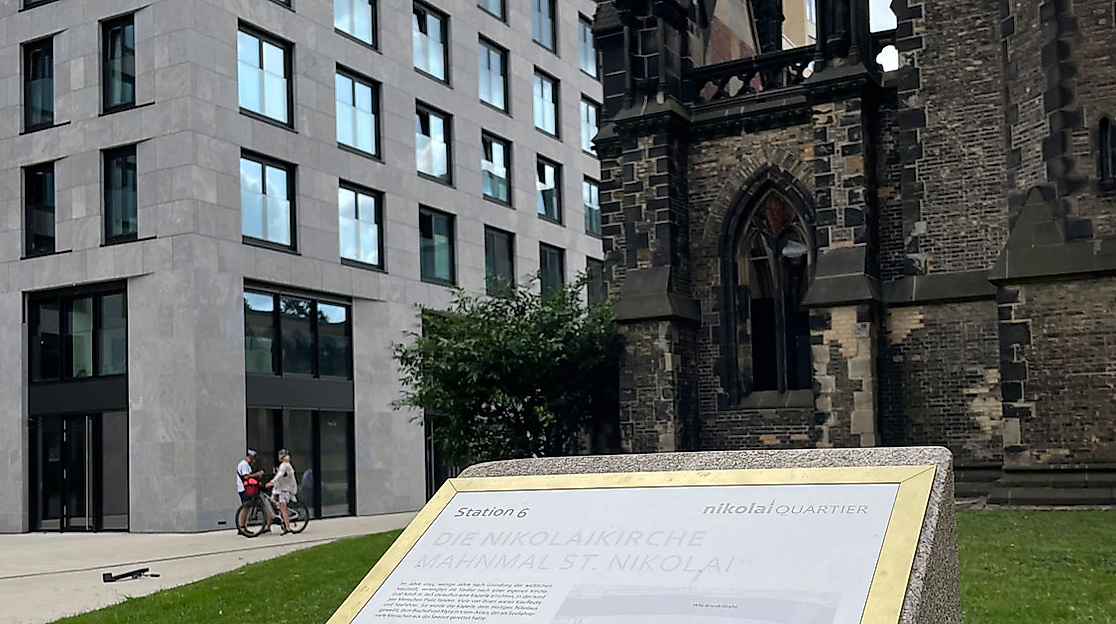 Infotafel vor der Ruine St. Nikolai in Hamburg mit modernem Gebäude im Hintergrund und grünem Vorplatz