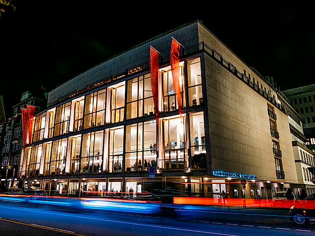 Beleuchtete Außenfassade der Hamburgischen Staatsoper bei Nacht mit Lichtspuren vorbeifahrender Autos.