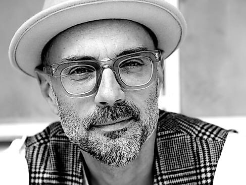Ein Foto eines Mannes mittleren Alters mit teilweise grauen Bartstoppeln und einem großen Brillengestell mit dickem, transparentem Rahmen. Er trägt einen hellen Fedora-Hut und eine karierte Weste.