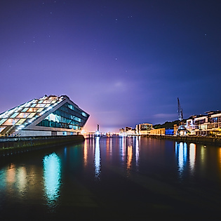Beleuchtetes Dockland-Gebäude bei Nacht mit Lichtspiegelungen im Wasser der Elbe und Hafenpanorama