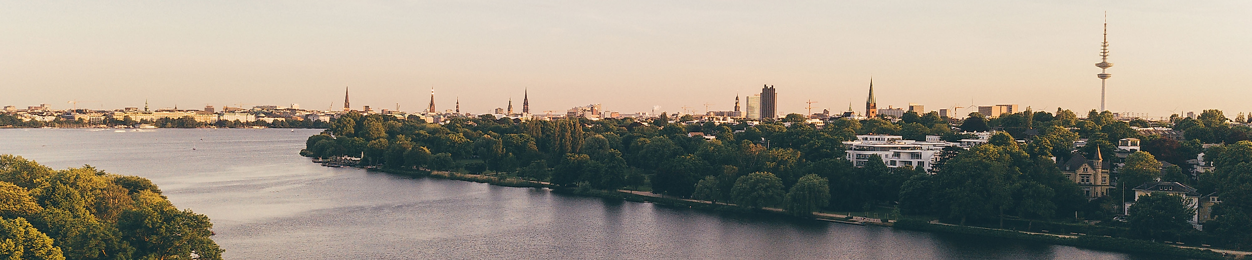 Luftaufnahme der Außenalster mit Segelbooten und Blick auf Hamburgs Skyline im Abendlicht.
