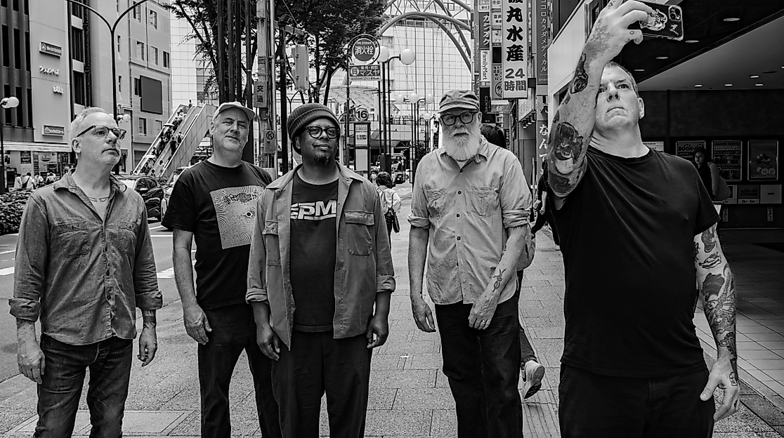 Fünf Personen stehen beieinander und blicken nach oben, eine Person hat das Handy hochgehalten und scheint ein Selfie von der Gruppe zu machen. Sie stehen inmitten einer asiatischen Großstadt.