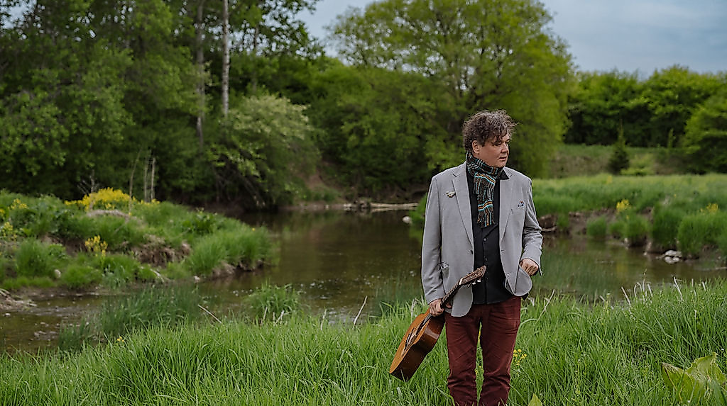 Ron Sexsmith steht in einer grünen Landschaft und hält seine Gitarre in der Hand.