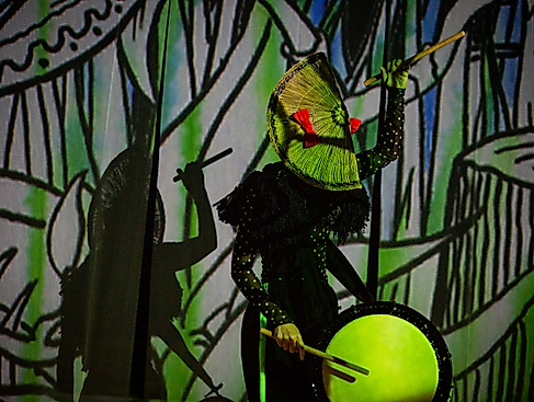 Eine Person trommelt vor einer in grün-schwarzen Symbolen angestrahlte Wand. Die eine Hand mit einem Drumstick in der Höhe, die andere an der Trommel. Auf dem Kopf sitzt eine fächerartige Kopfbedeckung.