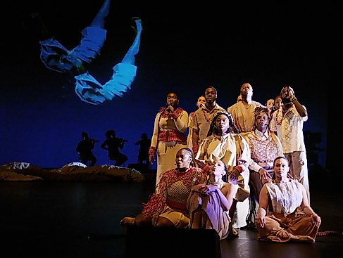 Eine Gruppe von Performerinnen in beigen und roten Kostümen sitzt und steht in einer Formation auf der Bühne. Im Hintergrund sind Silhouetten von Musikerinnen und eine Projektion eines schwebenden Körpers zu sehen.