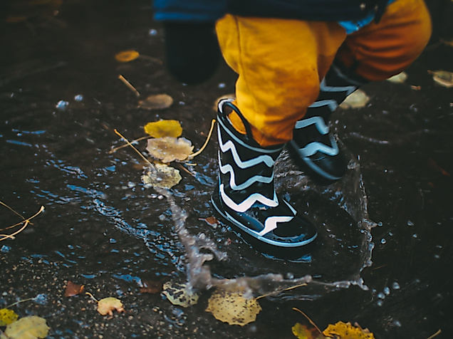 Kind springt mit gemusterten Gummistiefeln in eine Pfütze zwischen buntem Herbstlaub in Hamburg