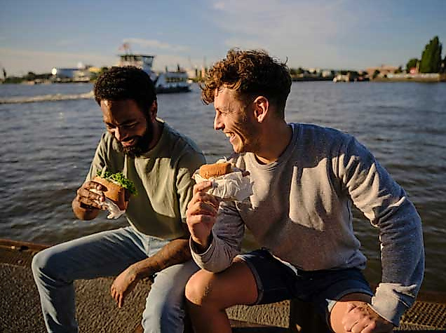 Zwei junge Männer sitzen entspannt am Wasserufer der Elbe in Hamburg und genießen Burger und Getränke im Sonnenlicht. Im Hintergrund ist ein Hafenboot auf dem Wasser zu sehen. Die Stimmung ist locker und sommerlich – ein typischer Genussmoment bei einem Picknick oder Snack mit Blick auf den Hafen, wie ihn Hamburg mit der Hamburg CARD erlebbar macht.