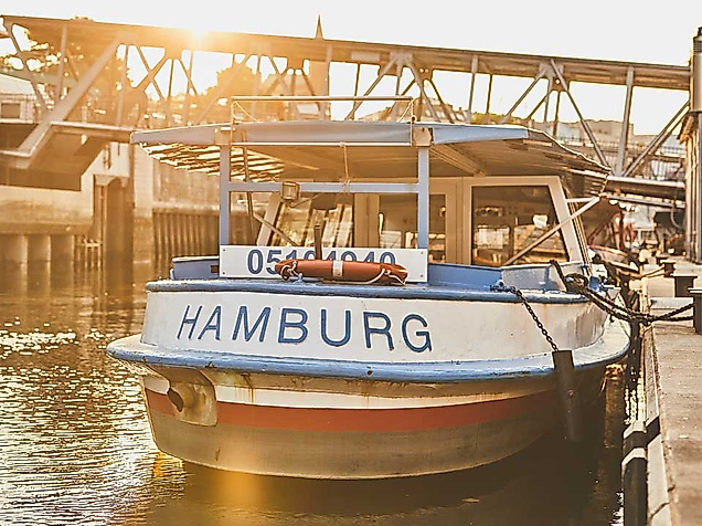 Ein traditionelles, blau-weißes Hafenrundfahrtboot mit der Aufschrift „HAMBURG“ liegt sonnenbeschienen an einem Anleger, das Wasser glitzert goldfarben. Über dem Boot spannt sich eine filigrane Stahlbrücke, im Hintergrund sind Backsteingebäude der Speicherstadt zu erahnen. Die Szene weckt Sehnsucht nach einer gemütlichen Barkassenfahrt – mit der Hamburg CARD sogar vergünstigt.