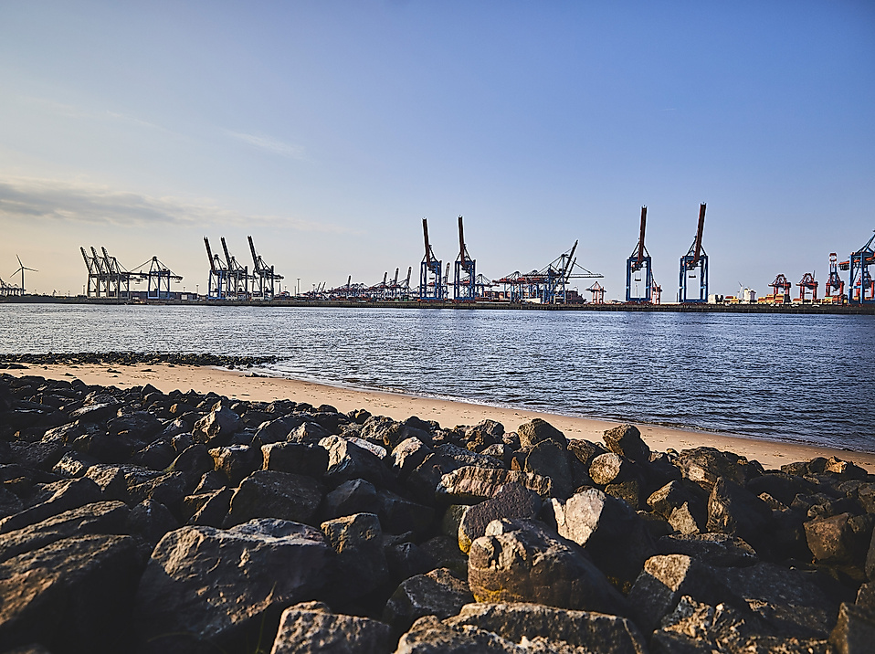 Blick vom Elbstrand auf Containerkräne im Hamburger Hafen bei Abendlicht, mit Steinen im Vordergrund