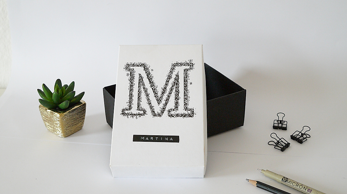 Lettering-Postkarte mit verziertem Buchstaben „M“, daneben Deko, Stifte und kleine Sukkulente auf hellem Tisch.