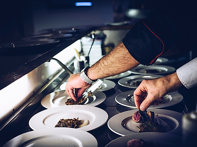Koch richtet kunstvoll kleine Portionen Fusionsküche auf mehreren weißen Tellern an.
