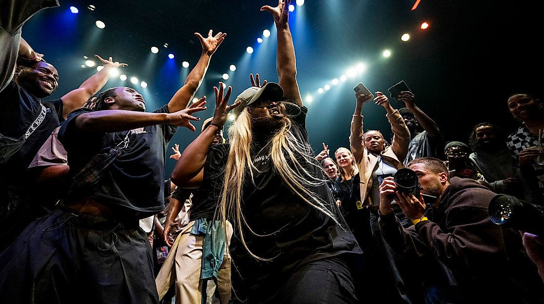 Eine dynamische Szene, bei der einen menge Menschen um eine Tänzerin herumstehen und sie mit Gesten anfeuern und jubeln. Die Tänzerin hebt streckt ihre Arme nach oben, spreizt die Finger. Rechts unten fotografiert jemand die Tänzerin von Nahem.
