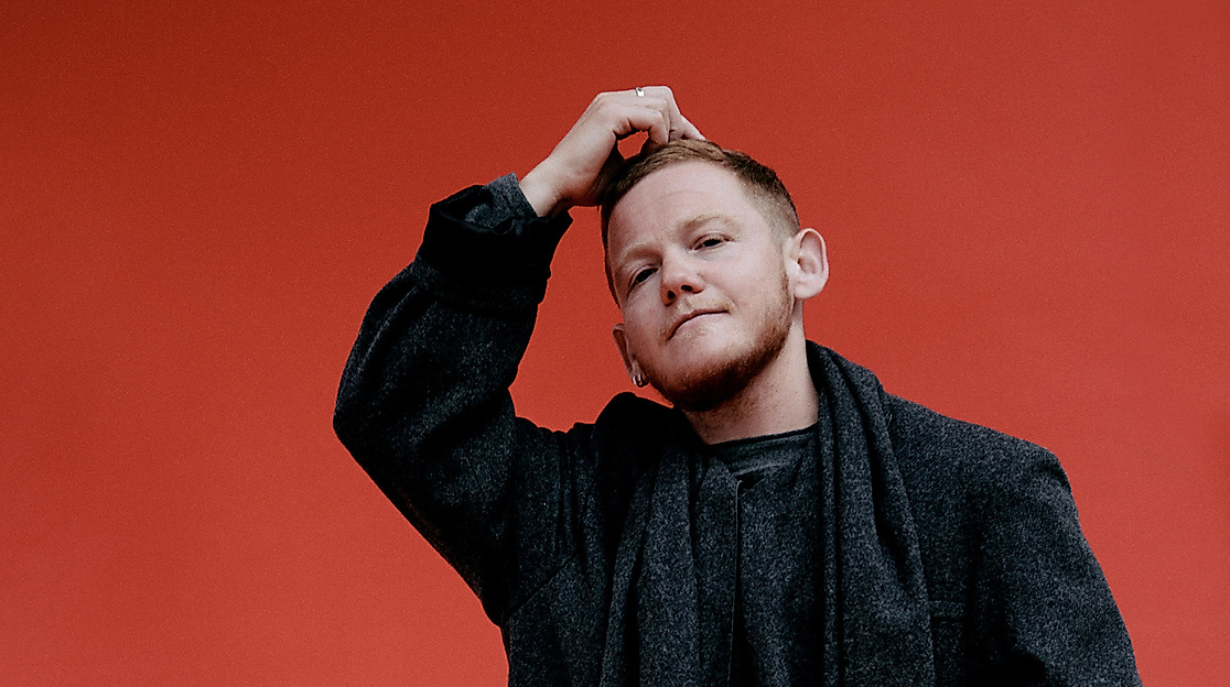 Kae Tempest steht in schwarz gekleidet vor einer bunten Wand und hat eine Hand auf den Kopf gelegt.