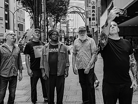 Fünf Personen stehen beieinander und blicken nach oben, eine Person hat das Handy hochgehalten und scheint ein Selfie von der Gruppe zu machen. Sie stehen inmitten einer asiatischen Großstadt.