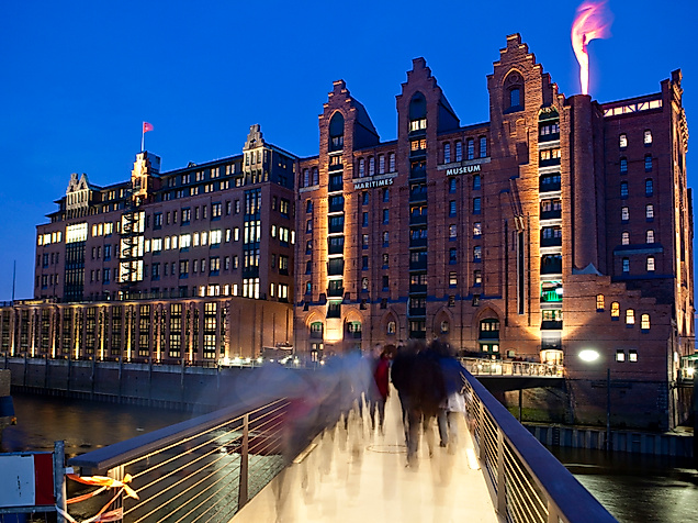 Internationales Maritimes Museum in der beleuchteten Speicherstadt am Abend mit Fußgängerbrücke im Vordergrund.