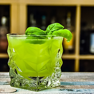 Grüner Cocktail mit Minze und Limettengarnitur im Glas vor einer Bar mit Flaschenregal im Hintergrund