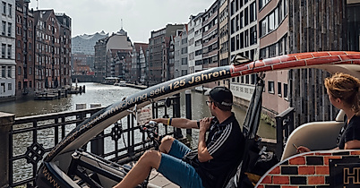 Zwei Personen fahren mit einer Rikscha durch die Hamburger Speicherstadt entlang eines Fleets.