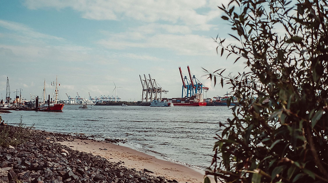 Blick vom Elbstrand Ottensen auf Schiffe und Containerkräne im Hamburger Hafen bei teils bewölktem Himmel.