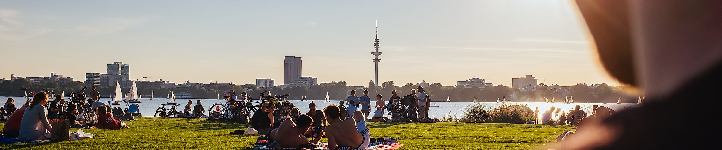 Menschen entspannen im Alsterpark bei Sonnenschein mit Blick auf die Außenalster und die Skyline Hamburgs