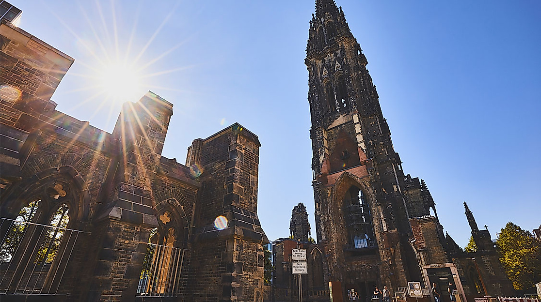Ruine der St.-Nikolai-Kirche in Hamburg bei Sonnenschein, mit Mahnmal und neugotischem Turm vor blauem Himmel
