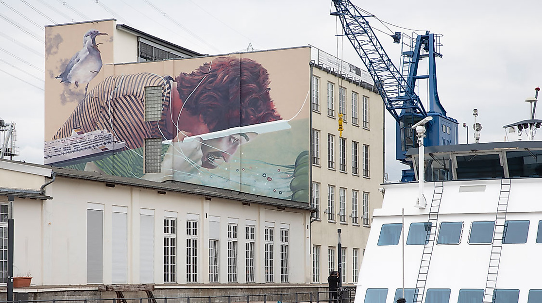 Fassadenkunst mit maritimem Motiv auf einem Hafengebäude in Hamburg-Harburg, daneben Kran und Schiff im Hafen