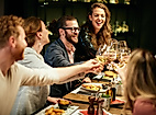 Gruppe junger Menschen lacht und stößt an einem lebhaften Restauranttisch mit Getränken an