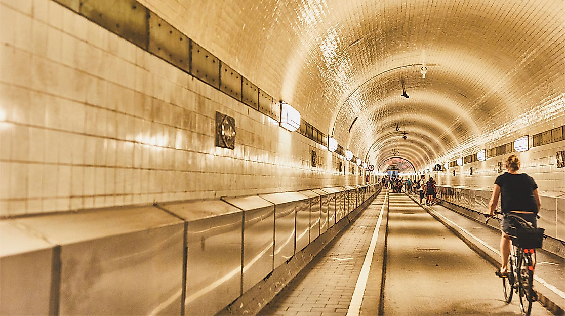 Fahrradfahrende und Fußgänger:innen im beleuchteten Alten Elbtunnel mit gekachelten Wänden in Hamburg