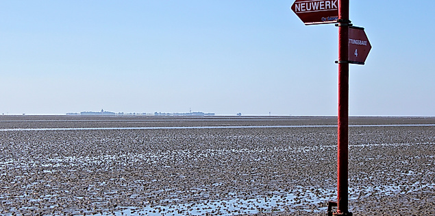 Wegweiser im Watt vor Neuwerk bei Ebbe, zeigt Richtung Cuxhaven und Insel, im Hintergrund Wattenmeer und Horizont.