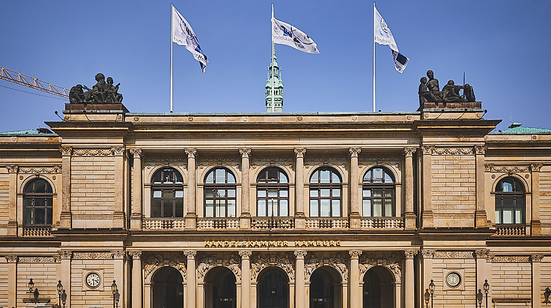 Fassade der Handelskammer Hamburg mit Flaggen, Rundbogenfenstern und klassizistischen Säulen vor blauem Himmel