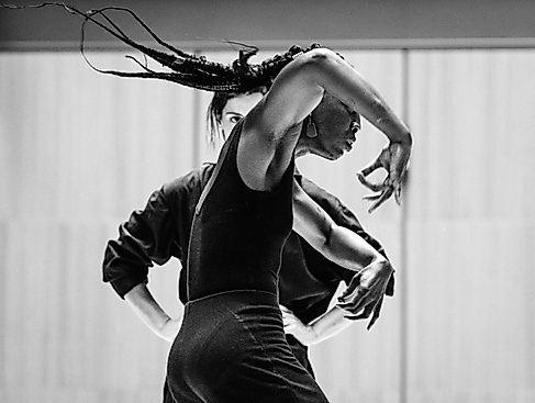 Eine Performerin ist von der Seite zu sehe, sie bewegt ihre Arme ausdrucksstark. Im Hintergund ist eine zweite Person zu sehen, die sie dabei beobachtet.