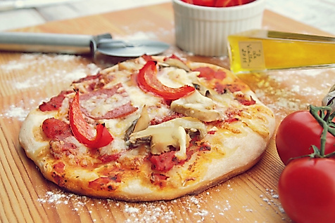 Frisch gebackene Pizza mit Paprika, Pilzen und Schinken auf Holzbrett, daneben Tomaten und Pizzaschneider.