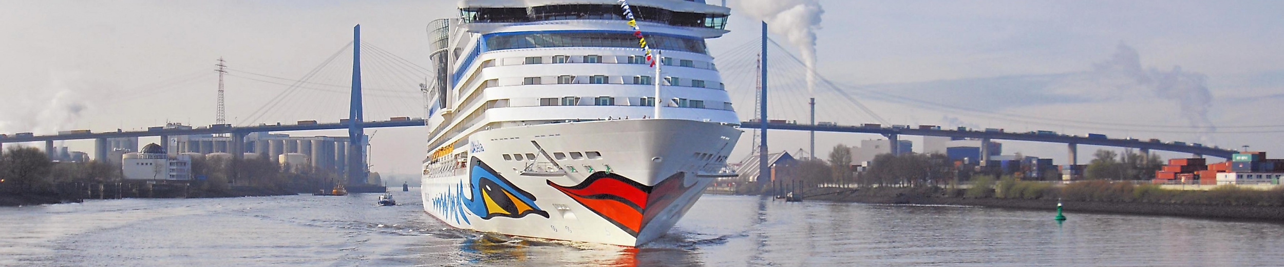 AIDA Bella fährt durch den Hamburger Hafen, im Hintergrund die Köhlbrandbrücke und Industrieanlagen
