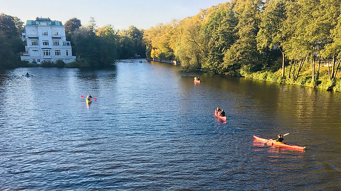 Menschen im Kajak auf einem Alsterkanal in Hamburg, umgeben von Bäumen und Villa im Abendlicht