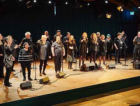 Eine Gruppe von Menschen steht auf einer Bühne und singt gemeinsam in Mikrofone.