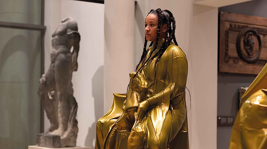 Eine Person in einen glänzenden goldfarbenen Anzug sitzt als Ausstellungsstück im Museum.