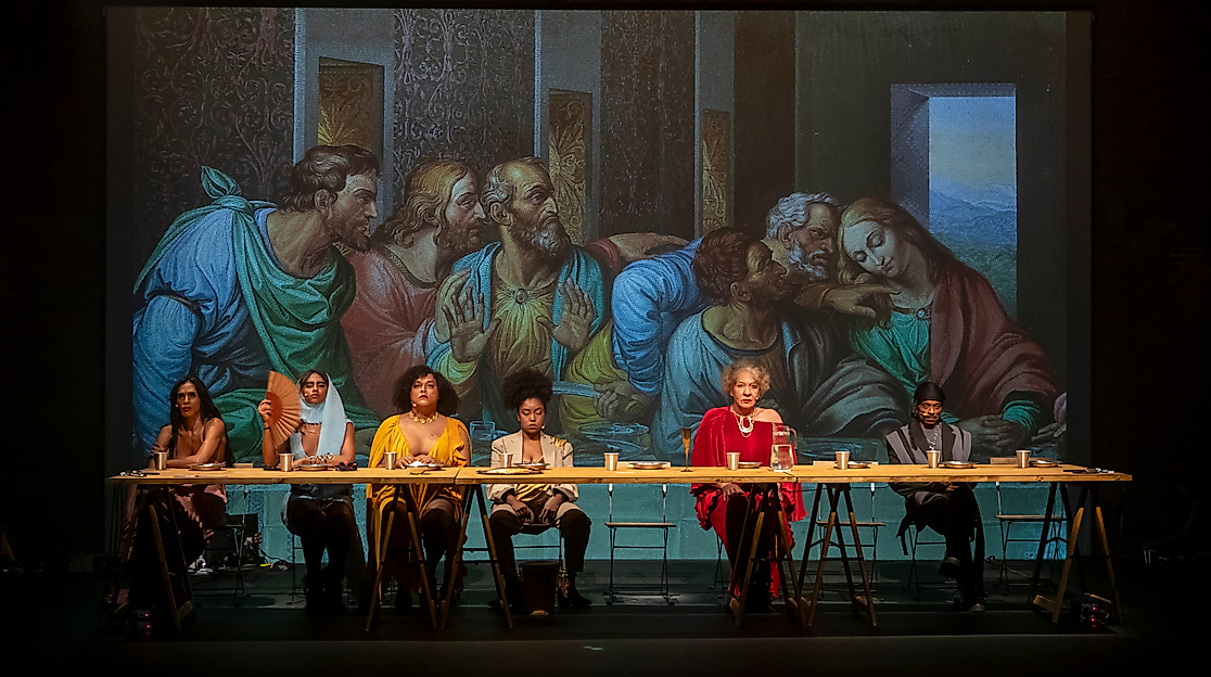 Performer*innen sitzen an einer langen Tafel vor einer Projektion von Da Vincis »Letztem Abendmahl« — eine zeitgenössische Inszenierung mit vielfältigem Ensemble.