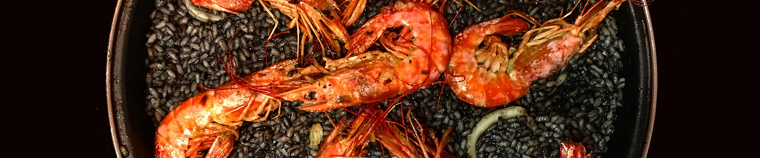 Pfanne mit schwarzer Paella und acht großen Garnelen auf dunklem Hintergrund stilvoll angerichtet.