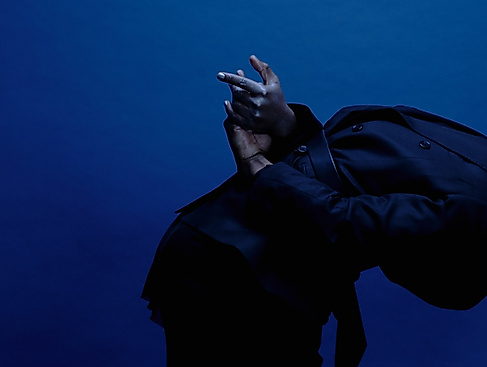 Ein*e Performer*in ist von der Seite zu sehen und steht nach vorne gebeugt, der schwarze Kaputzenpulli fällt über den Kopf. Die Hände sind hinter dem Rücken und ausgestreckt.