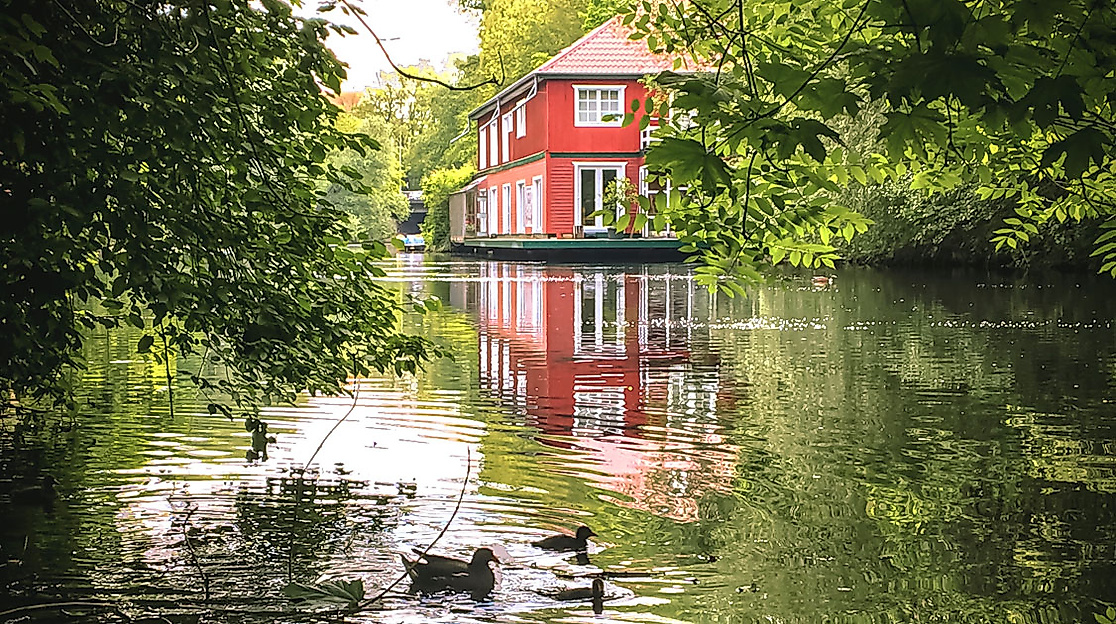 Rotes Haus am Alsterkanal, umgeben von grünen Bäumen und spiegelndem Wasser mit schwimmenden Enten