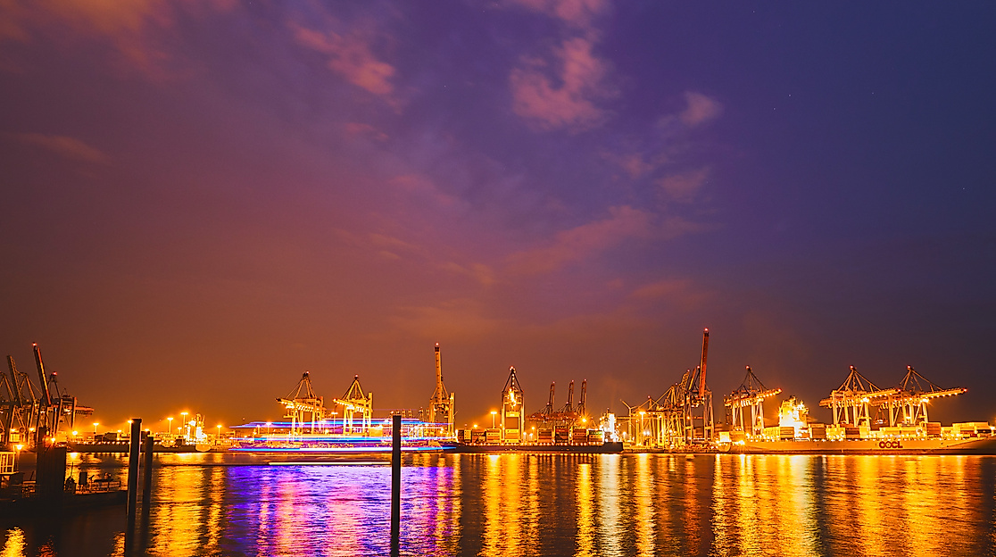 Beleuchtete Kräne und Schiffe im Hamburger Hafen bei Nacht, spiegelndes Wasser unter violettem Himmel