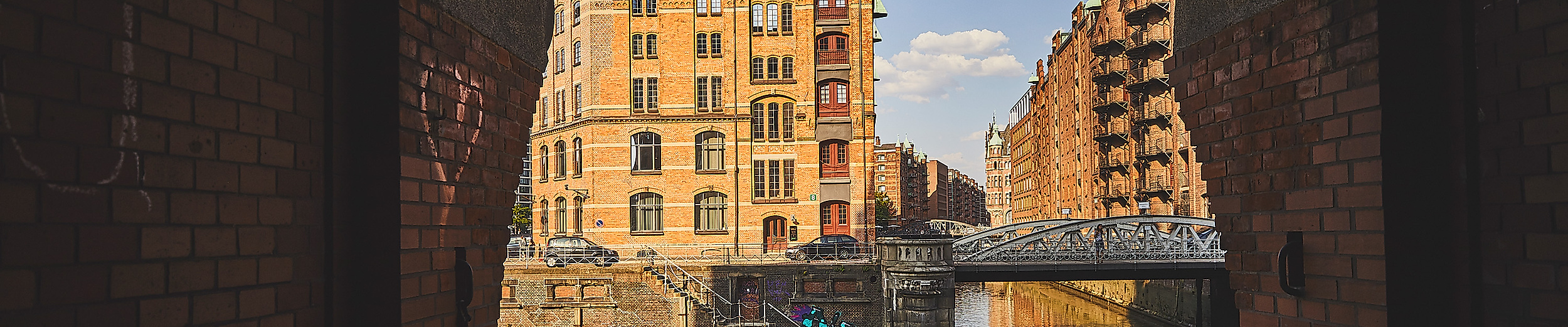 Blick durch einen Backsteinbogen in einen Innenhof der Speicherstadt mit Brücke und Wasserlauf bei Tageslicht