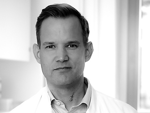Schwarz-weiß-Portraitfoto einer männlich gelesenen Person mittleren Alters mit großer Stirn und Segelohren. Sie trägt ein weißes Kragenhemd.