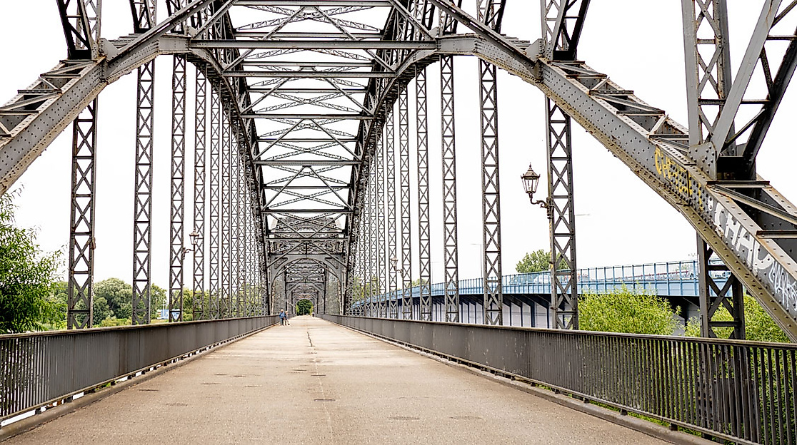 Historische Stahlbrücke mit Blickachse, umgeben von Bäumen, verbindet Ufer in Hamburg bei klarem Tageslicht