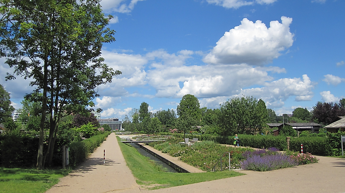 Breiter Weg mit Wasserlauf und Blumenbeeten im Wilhelmsburger Sommerpark unter blauem Himmel